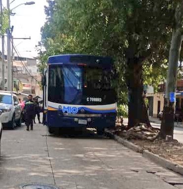 A blue bus with MIO branding is positioned across a street in a residential area lined with trees and buildings blocking the road with its front end crashed into a curb and dirt pile nearby. Two people in dark uniforms stand near the rear of the bus. A white car is parked adjacent to the scene under streetlights and power lines. The setting appears to be daytime in an urban neighborhood.