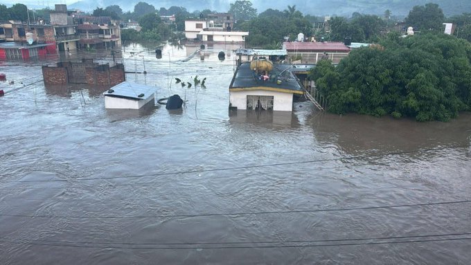 Floodwaters surround several low-rise buildings in a residential neighborhood, with some structures partially submerged and others elevated on stilts. Debris floats in the murky water, and green trees stand nearby. A few people are visible on rooftops or wading through the water, indicating a recent heavy rain event causing widespread inundation.