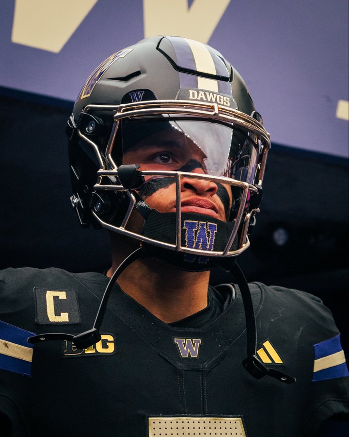 African American male football player in black helmet with purple stripes and DAWGS text, gold facemask, black jersey with white C on shoulder, W emblem, and purple accents, standing in front of purple wall with white W logo, expression intense with focused gaze.