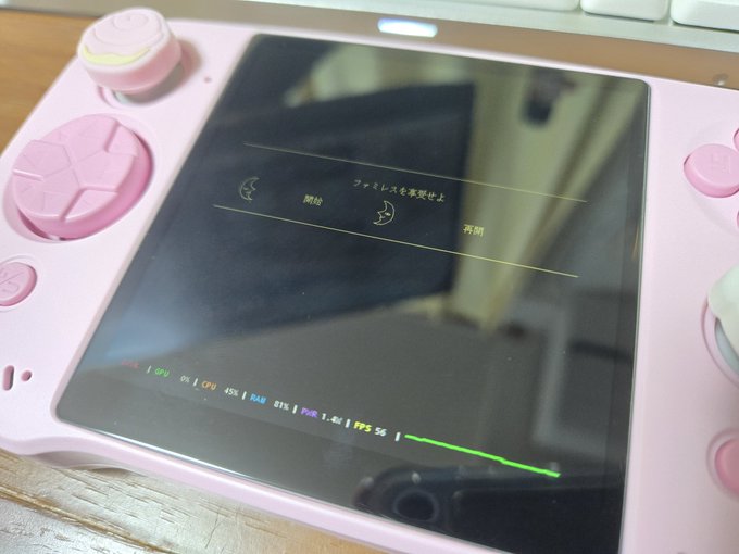Pink handheld gaming device with hexagonal pink buttons and pink rubber grips on a wooden desk next to a white keyboard, screen displaying game interface with Japanese text including Famiresu and menu options like sleep mode, compatibility layer details such as Proton9.0-x64-3, DXVK version dxvk-0.96, VKD3D None, and CPU translator Box64-0.37-b1, progress bar at bottom.