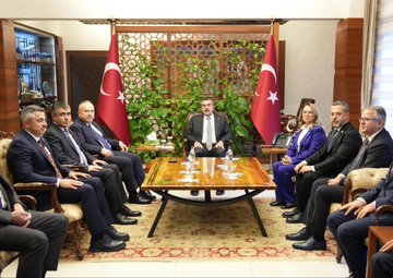 First image shows two men in dark suits standing at a wooden podium with Turkish flags on either side in a grand hall with ornate walls and lighting the podium labeled with gold text indicating governors office. Second image depicts four individuals two men and two women in formal suits and blazers posing together in front of Turkish flags in a similar ornate hall with geometric wall designs. Third image features a group of eight people including men and women in business attire seated around a wooden table with plants and water glasses in a room with wooden paneling and more Turkish flags.