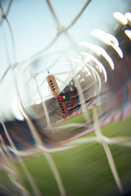 A soccer goalpost with a net in the foreground, viewed from behind. A stadium field with green grass is visible through the net. In the background, a tall structure with a clock and colorful banners stands, surrounded by stadium seating and lights.