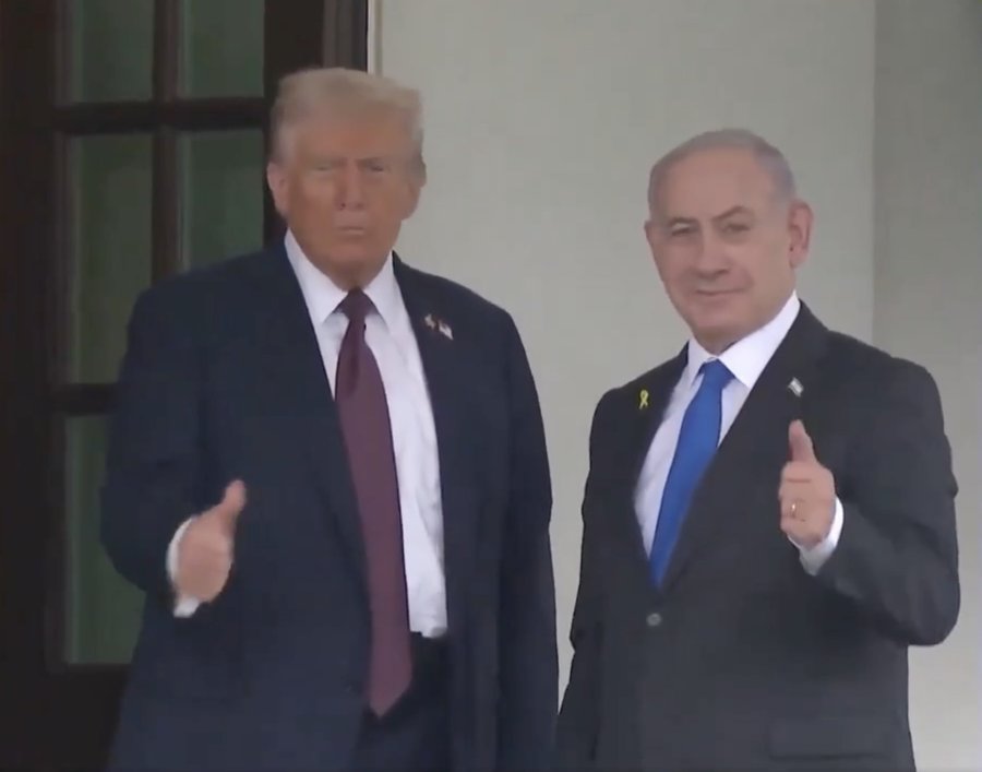 Donald Trump and Benjamin Netanyahu standing side by side, both wearing dark suits, white shirts, and ties. Donald Trump has a red tie and gives a thumbs-up gesture with his right hand. Benjamin Netanyahu has a blue tie and gives a thumbs-up gesture with his right hand. They are positioned in front of a building with large windows and white walls.