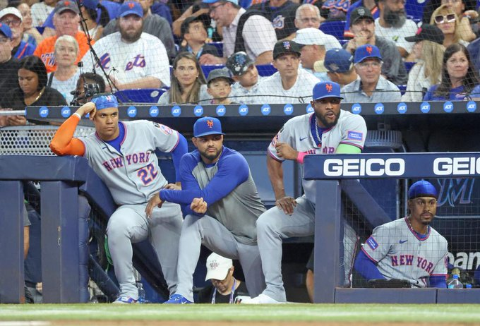 Four New York Mets players in gray uniforms with blue and orange accents, sitting in a dugout. Juan Soto is visible, wearing a cap and uniform with the number 22. The players are positioned near a railing with a GEICO advertisement. The crowd in the background is wearing Mets gear.