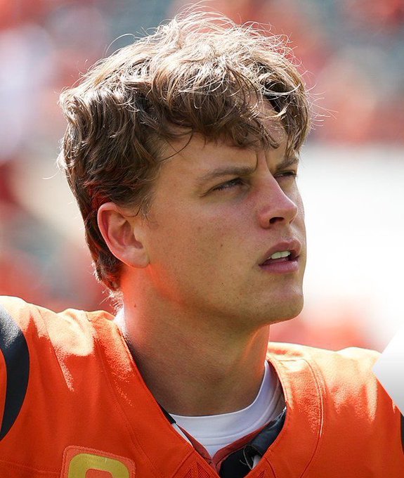 Joe Burrow wearing an orange football jersey with black and white accents, standing in a stadium setting with a blurred crowd in the background.