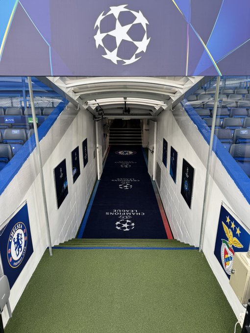 A tunnel leading to a soccer field, with blue and white walls. Chelsea FC logos are on the left wall, and Benfica logos are on the right wall. The floor has a blue carpet with "UEFA Champions League" text and the Champions League logo. Steps lead up to a green field, with stadium seating visible on both sides.