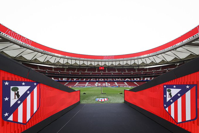 A view of the Wanda Metropolitano stadium, home of Atlético Madrid. The image shows a tunnel leading to the field, flanked by red walls with the Atlético Madrid logo featuring red and white stripes. The field and stands are visible in the background, with the club's name and emblem displayed prominently.