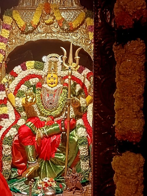 A statue of Durga Devi adorned with vibrant red and green garments, holding a trident, and decorated with garlands of flowers and jewelry. The statue is framed by an ornate arch with yellow and white floral arrangements. Marigold garlands are visible on the sides of the frame.