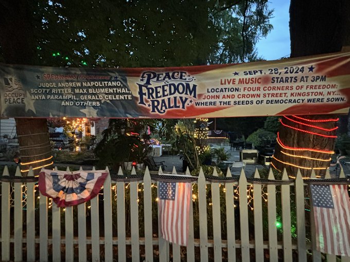 A banner reading "Peace & Freedom Rally" with text stating "Sept. 28, 2024" and "Live Music Starts at 4PM." The banner lists names including Judge Andrew Napolitano, Scott Ritter, Max Blumenthal, and others. American flags hang on a white picket fence below the banner, with string lights wrapped around trees. The setting includes a street view with houses and trees, visible during twilight.