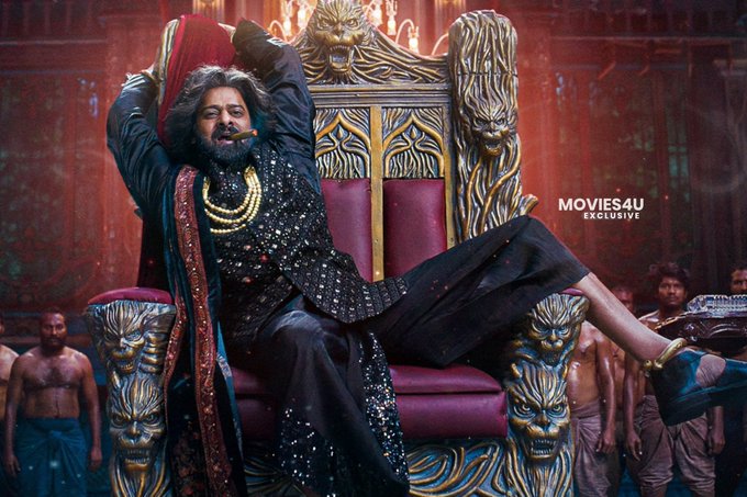 Prabhas sitting on an ornate throne with lion carvings and red cushions. He wears a black sequined outfit, a gold necklace, and a red sash, with one leg raised and an arm extended. Several muscular men in loincloths stand in the background. A watermark reading "MOVIES4U EXCLUSIVE" is visible in the top right corner.