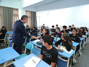 A man in a suit shaking hands with a student in a classroom filled with students at desks, wearing casual clothing. Another image shows the same man in a suit among a group of people, including students and adults, standing outdoors in front of a building with a sign reading "Ankara-Çankaya Betül Can Anadolu Lisesi." A third image depicts the same group posing for a photo outdoors in front of the same building.