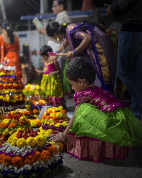 Women and children in colorful traditional attire arranging vibrant floral arrangements in conical shapes. The flowers include marigolds, roses, and other blossoms, adorned with lit candles. A statue of a goddess decorated with flowers and a candle is visible, surrounded by similar floral displays.