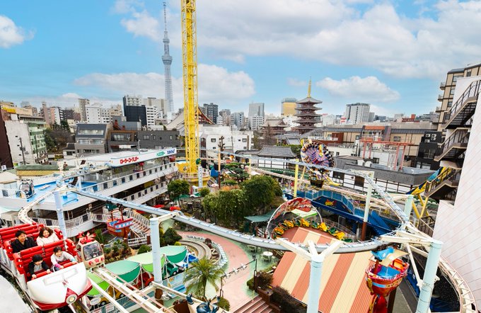 A vibrant amusement park with colorful rollercoasters, rides, and structures. A yellow tall ride and a red ride with people are visible. The Tokyo Skytree and a pagoda are seen in the background skyline.