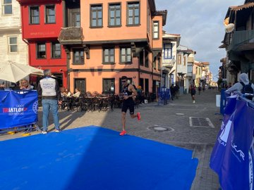 A group of athletes in red and orange uniforms standing on a blue platform, holding medals and a banner reading "TSK Spor Gücü Komutanlığı." They are under a blue arch with text "Europe Balkan Championships 2025 Mudanya" and logos. Trophies are placed in front. An athlete runs on a blue mat in a street with colorful buildings. Two cyclists in black and red outfits ride on a road near palm trees and buildings. A man in a military uniform holds a trophy on a podium, surrounded by people in suits, with horses visible in the background.