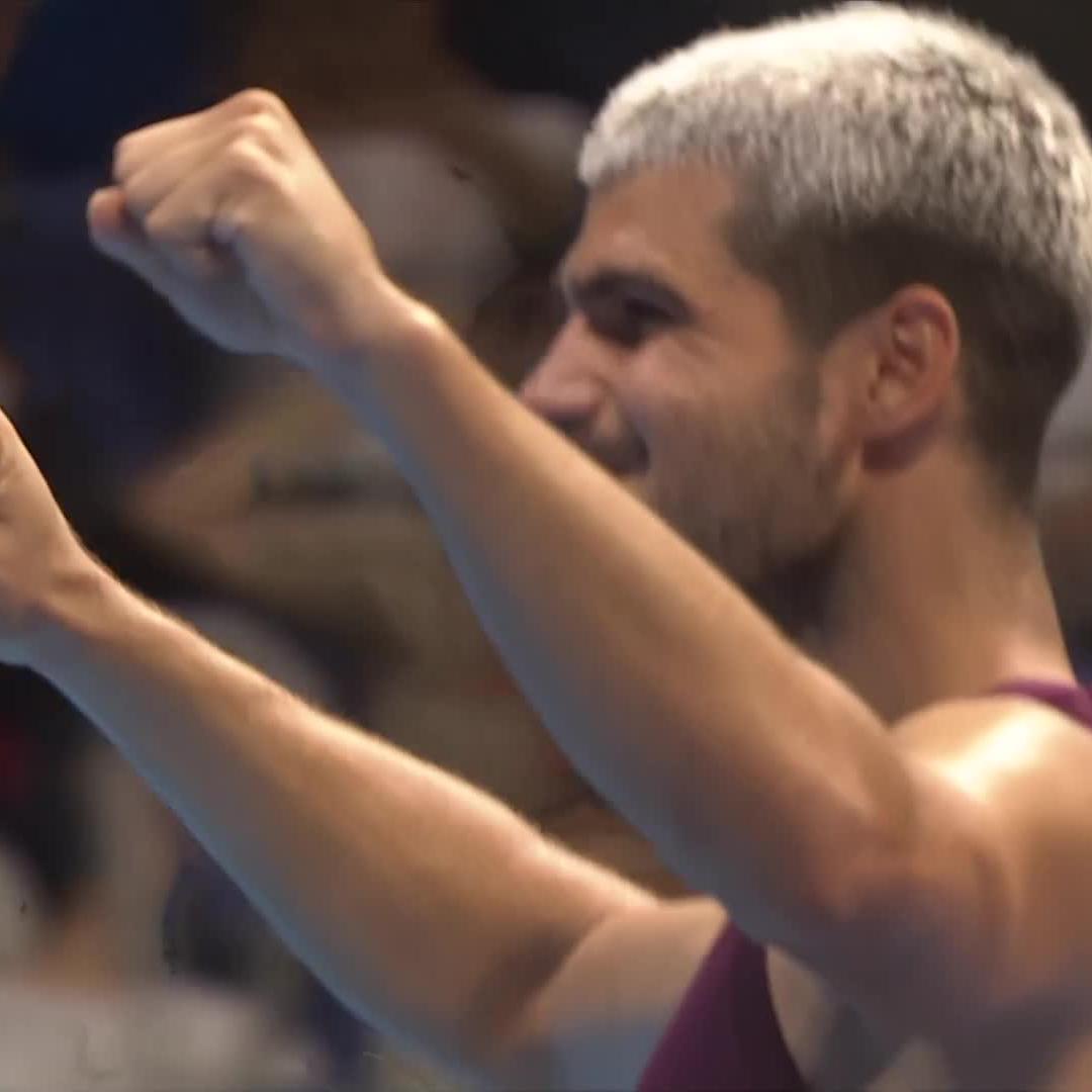 Carlos Alcaraz, the new champion of the Tokyo 2025 tournament, raises his fists in celebration while wearing a maroon sleeveless shirt, with his signature bleached blonde hair now grown out to a darker shade—earning him the fan nickname "Blondecaraz" that supporters say they miss.