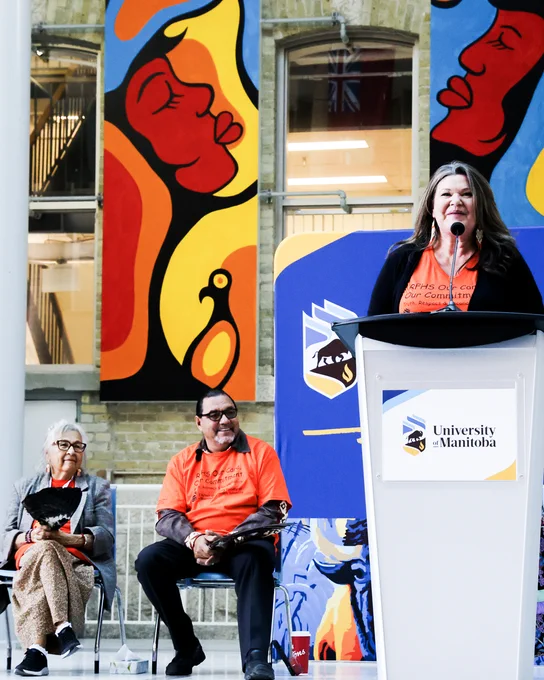 A close-up shot of a person with a confident, focused expression, with long, dark hair hanging over their shoulders, wearing an orange shirt under an open, dark jacket, standing behind a podium. Behind, two others are seated, watching, wearing orange shirts.