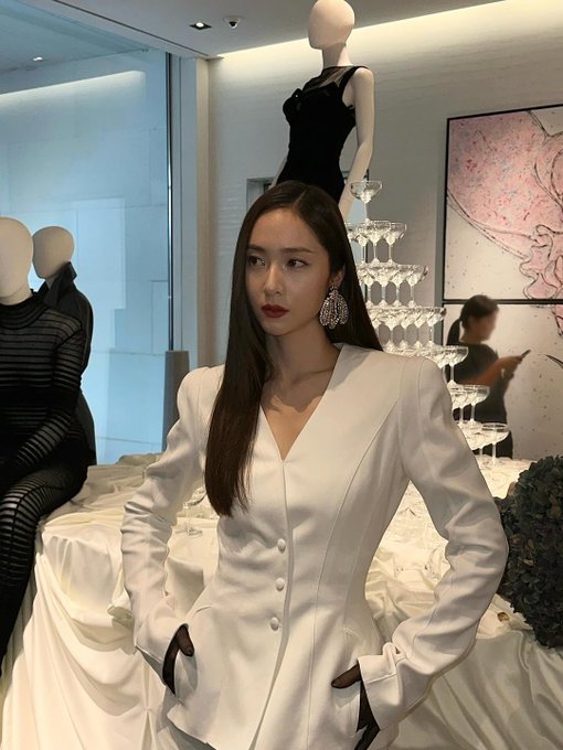 Krystal Jung stands in a stylish white blazer with long sleeves and black gloves, paired with black pants. Her long, straight brown hair falls over her shoulders, and she wears large, sparkling earrings. Mannequins dressed in black and white outfits are positioned nearby, displaying similar fashion styles. The setting includes a modern interior with white tables, glassware, and framed artwork on the walls.
