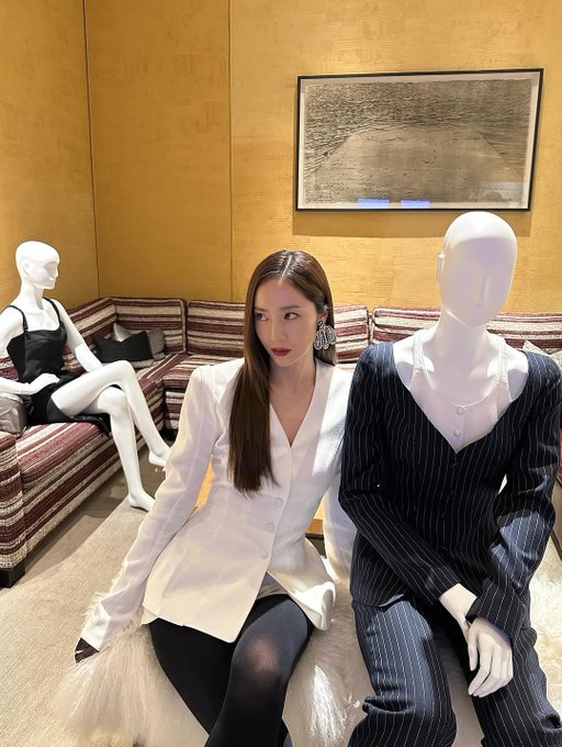 Krystal Jung stands in a stylish white blazer with long sleeves and black gloves, paired with black pants. Her long, straight brown hair falls over her shoulders, and she wears large, sparkling earrings. Mannequins dressed in black and white outfits are positioned nearby, displaying similar fashion styles. The setting includes a modern interior with white tables, glassware, and framed artwork on the walls.