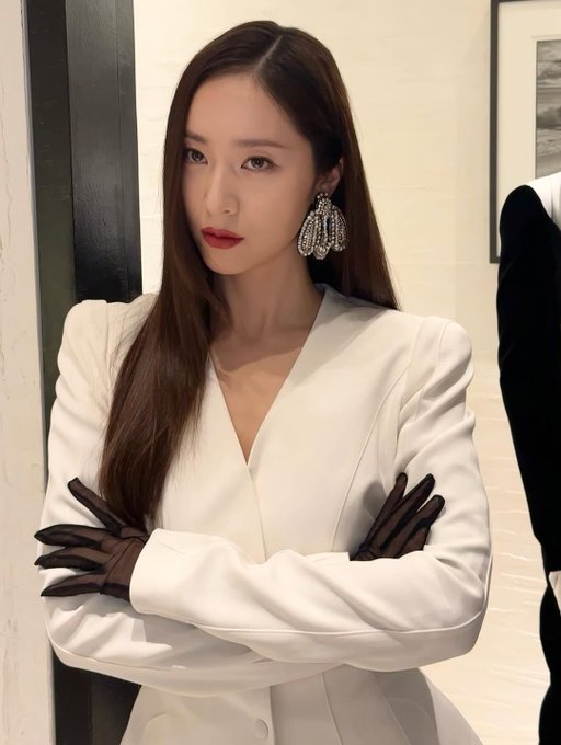 Krystal Jung stands in a stylish white blazer with long sleeves and black gloves, paired with black pants. Her long, straight brown hair falls over her shoulders, and she wears large, sparkling earrings. Mannequins dressed in black and white outfits are positioned nearby, displaying similar fashion styles. The setting includes a modern interior with white tables, glassware, and framed artwork on the walls.