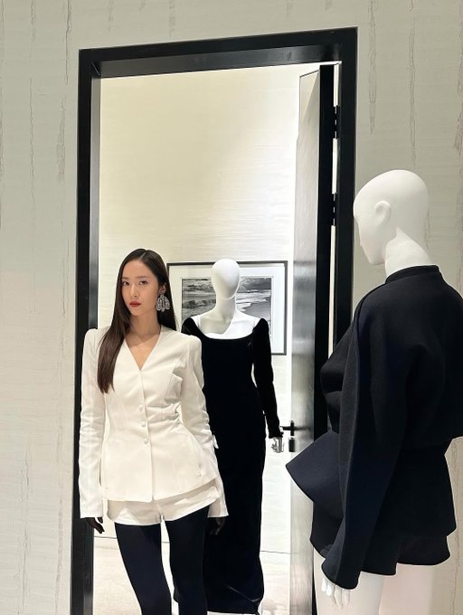 Krystal Jung stands in a stylish white blazer with long sleeves and black gloves, paired with black pants. Her long, straight brown hair falls over her shoulders, and she wears large, sparkling earrings. Mannequins dressed in black and white outfits are positioned nearby, displaying similar fashion styles. The setting includes a modern interior with white tables, glassware, and framed artwork on the walls.