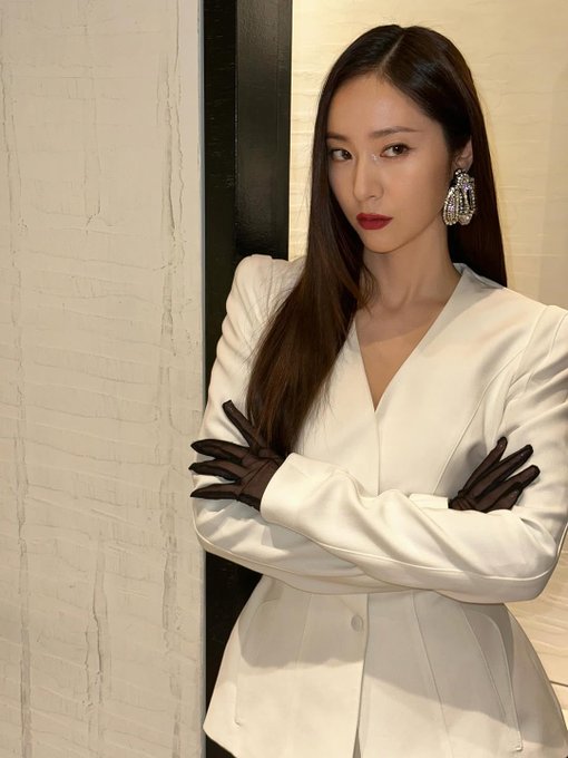Krystal Jung wearing a white tailored blazer with long sleeves and a V-neck, paired with black gloves. Her long brown hair is straight and falls over her shoulders. She stands with arms crossed, wearing large, sparkling earrings.