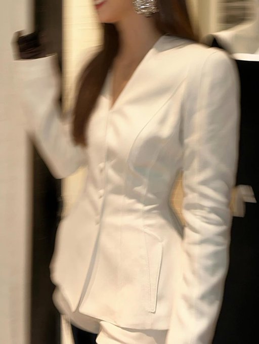 Krystal Jung wearing a white tailored blazer with long sleeves and a V-neck, paired with black gloves. Her long brown hair is straight and falls over her shoulders. She stands with arms crossed, wearing large, sparkling earrings.
