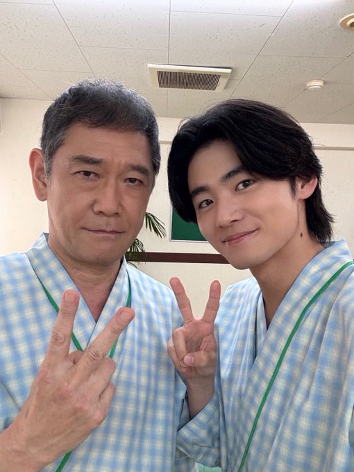 Two men, Haruhi Iuchi and another individual, wearing matching light blue checkered hospital gowns with green trim, making peace signs with their hands. Another image shows two men, Haruhi Iuchi and another individual, in casual attire, one in a beige suit and white shirt, the other in a black shirt and blue jeans, both making peace signs. A third image displays a wrapped gift box with Japanese text and colorful mask illustrations. No watermarks are present.