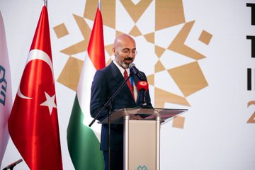 Mahmut Özdil speaking at a podium with Turkish and Hungarian flags, microphones, and a backdrop with geometric patterns. A group including Numan Kurtulmuş, Laszlo Köver, Fatih Dönmez, and Gülşen Karanis standing together cutting a red ribbon at a ceremony. Mahmut Özdil addressing an audience at a podium with flags and a backdrop featuring text "TÜRKİYE 2023". Mahmut Özdil and others standing with students in a classroom setting with desks and educational materials.
