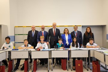 Mahmut Özdil speaking at a podium with Turkish and Hungarian flags, microphones, and a backdrop with geometric patterns. A group including Numan Kurtulmuş, Laszlo Köver, Fatih Dönmez, and Gülşen Karanis standing together cutting a red ribbon at a ceremony. Mahmut Özdil addressing an audience at a podium with flags and a backdrop featuring text "TÜRKİYE 2023". Mahmut Özdil and others standing with students in a classroom setting with desks and educational materials.