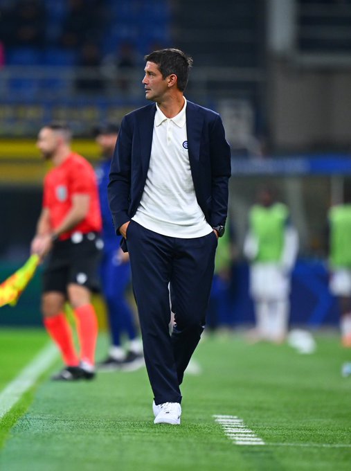 A man wearing a white polo shirt, dark blazer, dark pants, and white sneakers stands on a soccer field. He has short brown hair and is looking to the side. In the background, a referee in a red shirt and black shorts, and other individuals in green and white training kits are visible on the field.