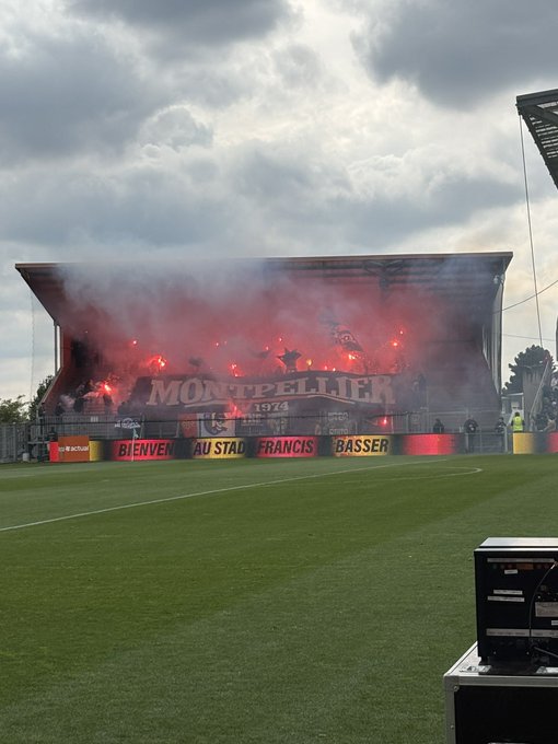 A soccer field with a grandstand in the background. Fans stand in the grandstand holding banners and flares, with smoke rising. The banner reads "MONTPELLIER 1974" and a sign below says "BIENVENUE AU STADE FRANÇOIS COTY LE BASSE." The field is green with white lines, and the sky is cloudy.