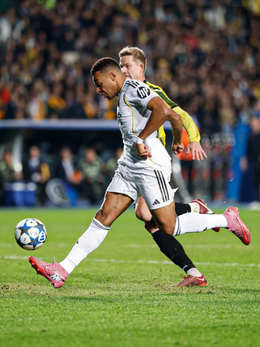 Kylian Mbappé in a white Real Madrid jersey with black and gold accents, kicking a soccer ball on a grassy field. He wears pink cleats and white socks. An opposing player in a yellow and black jersey attempts to block him. The Champions League ball is visible under Mbappé\'s foot.