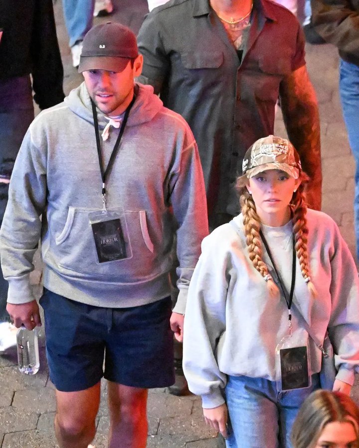 Sydney Sweeney wearing a camouflage cap and light gray sweatshirt with braids, holding lanyards. Scooter Braun in a gray hoodie, cap, and shorts, also holding lanyards. Sydney Sweeney\'s parents, a woman in a black top and jeans and a man in a gray shirt, walking together at Universal Studios Hollywood. Lanyards display badges with text.