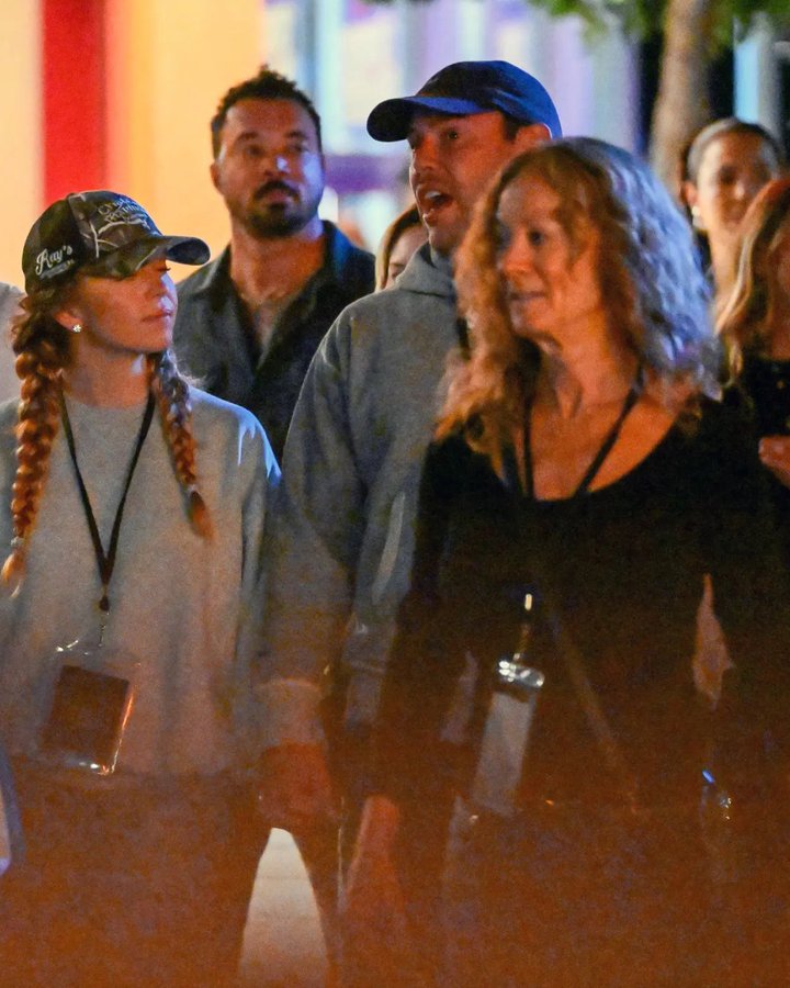 Sydney Sweeney wearing a camouflage cap and light gray sweatshirt with braids, holding lanyards. Scooter Braun in a gray hoodie, cap, and shorts, also holding lanyards. Sydney Sweeney\'s parents, a woman in a black top and jeans and a man in a gray shirt, walking together at Universal Studios Hollywood. Lanyards display badges with text.