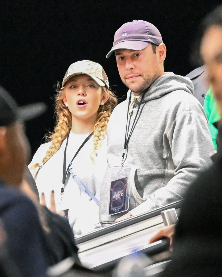 Sydney Sweeney wearing a camouflage cap and light gray sweatshirt with braids, holding lanyards. Scooter Braun in a gray hoodie, cap, and shorts, also holding lanyards. Sydney Sweeney\'s parents, a woman in a black top and jeans and a man in a gray shirt, walking together at Universal Studios Hollywood. Lanyards display badges with text.