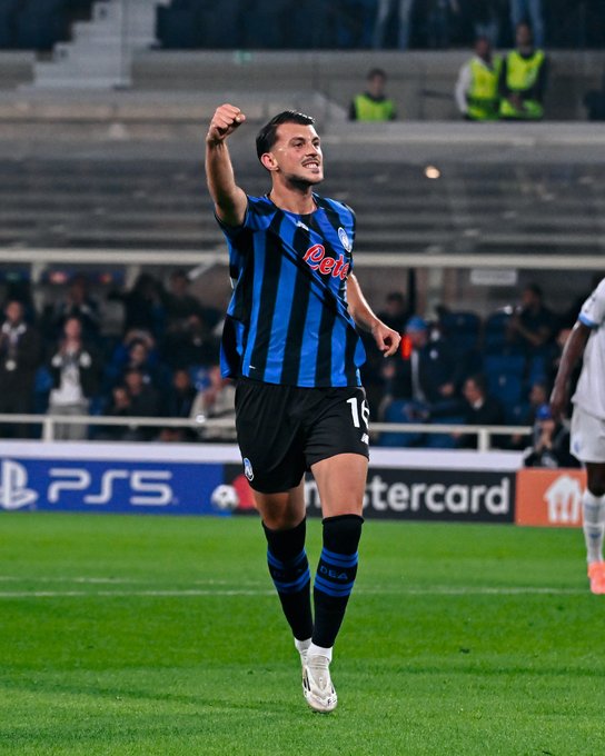 A soccer player wearing a black and blue striped Atalanta jersey with the number 16, raising one arm in celebration on a grassy field. The player has dark hair and is wearing black shorts and black socks with blue stripes. Spectators and stadium seating are visible in the background, along with advertising boards displaying "PlayStation 5" and "Mastercard".