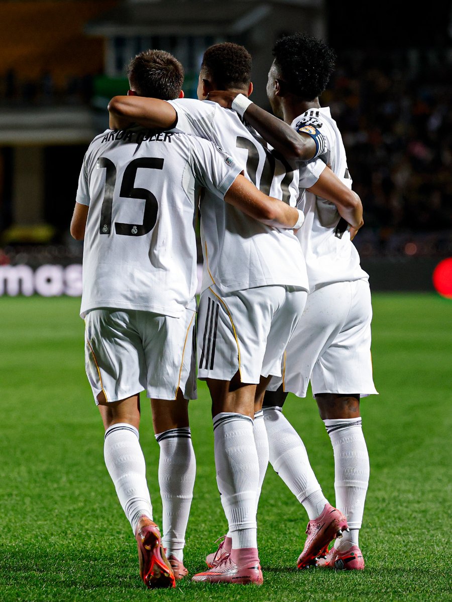 Arda Güler, Kylian Mbappé, and Vinícius Jr. wearing white Real Madrid jerseys with black and gold accents, standing on a grassy field with their arms around each other. Their jersey numbers are visible: 15 on Arda Güler, 9 on Kylian Mbappé, and 7 on Vinícius Jr. They are wearing white socks and red cleats.