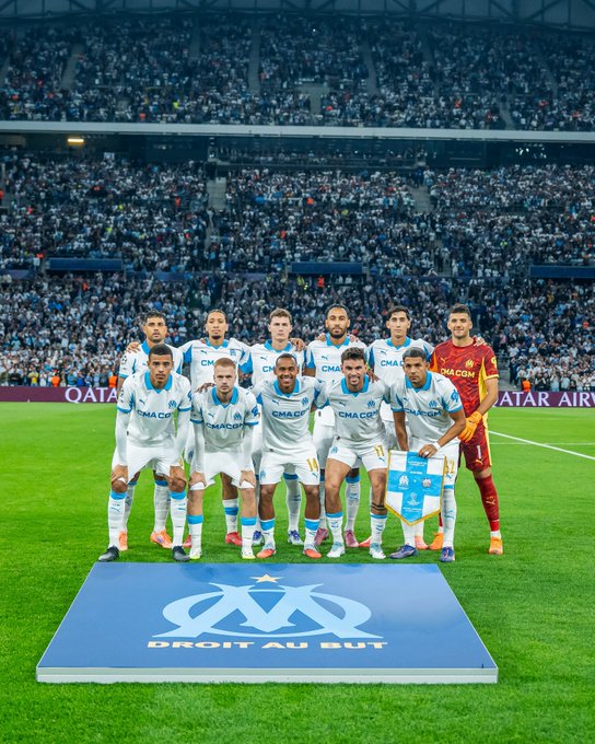 A group of soccer players from Olympique de Marseille wearing white and blue uniforms, standing on a field. They are positioned on a blue platform with the Olympique de Marseille logo and text "DROIT AU BUT." A goalkeeper in a red uniform stands to the right. A large crowd fills the stadium seats in the background.