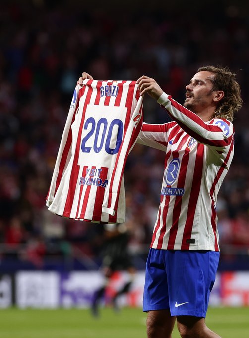 A soccer player wearing a red and white striped jersey with blue shorts holds up a matching jersey displaying the number 200. The jersey has the Atlético Madrid logo and text. The player stands on a grassy field with a crowd in the background.