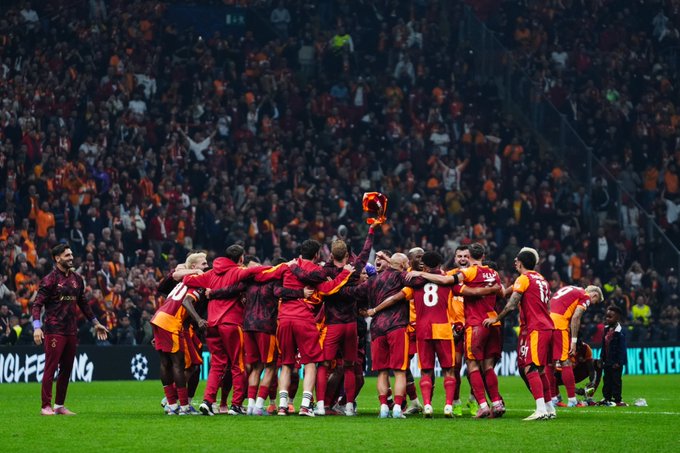 Galatasaray players in red and yellow uniforms huddle together on a soccer field, celebrating with arms around each other. One player holds a trophy above their heads. A large crowd of fans in the stands wears similar colors, filling the stadium.