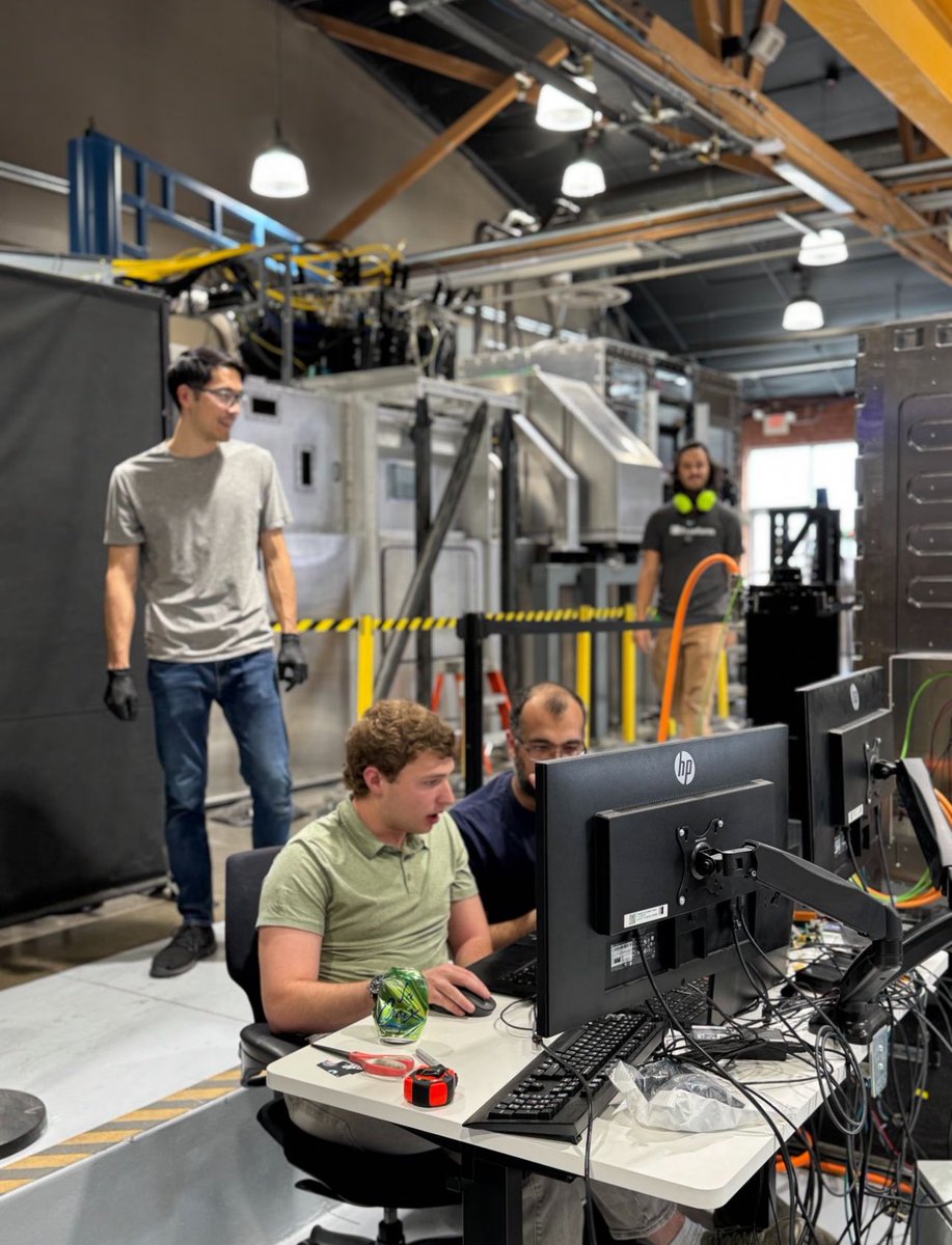ErikPalitsch's tweet photo. Engineering at its finest. Super proud of the work this team is doing everyday. 

Foreground: ME's, EE's, and SWE's deving out critical subsystems for our next-gen factory tech.

Background: The world's highest throughput metal printing system - producing perfect parts around the clock, with no human in the loop.