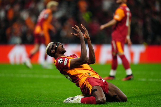 A soccer player in a red and yellow Galatasaray jersey kneels on a grassy field, raising both arms in celebration. The jersey displays the UEFA Champions League patch. Another player in a similar uniform stands in the background, with a blurred crowd visible beyond the field.