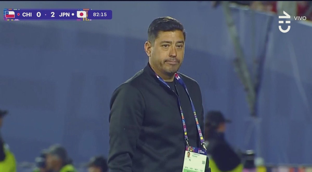 A man wearing a black jacket and lanyard with a badge, standing on a soccer field. A scoreboard shows CHI 0 - 2 JPN, with flags of Chile and Japan, and a time of 82:15. A Vivo watermark is visible in the corner.
