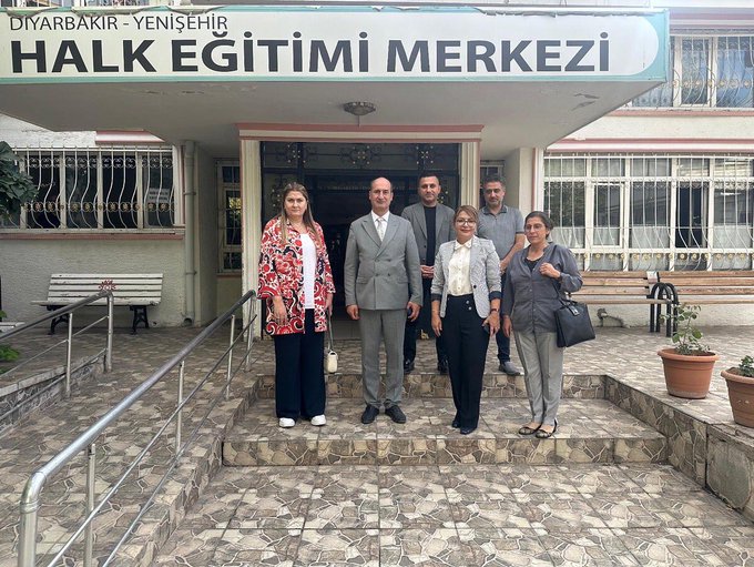 A group of people standing in front of a building with a sign reading "Olgunlaşma Enstitüsü Diyarbakır" and "Diyarbakır Yenişehir Halk Eğitimi Merkezi." The individuals are dressed in formal and semi-formal attire, including suits and dresses. The building has a pink exterior, windows, and potted plants near the entrance.