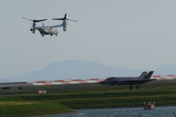 Four men in suits standing by water, looking at aircraft. A CMV-22 Osprey hovers in the sky above. An F-35B jet is visible on a runway in the distance, with mountains and a clear sky in the background.