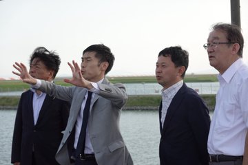 Four men in suits standing by water, looking at aircraft. A CMV-22 Osprey hovers in the sky above. An F-35B jet is visible on a runway in the distance, with mountains and a clear sky in the background.