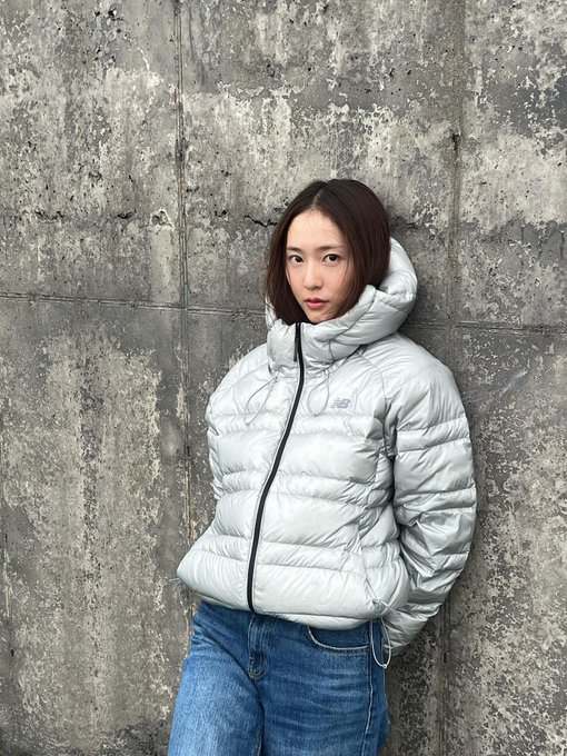 A person wearing a light gray New Balance puffer jacket with a hood and blue jeans, standing against a concrete wall. The same person, also in the New Balance jacket and jeans, stands among large rocks and greenery, wearing a gray beanie.