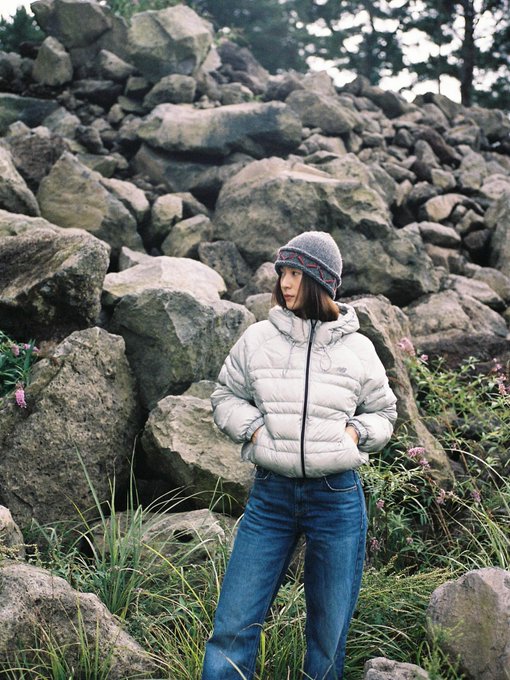 A person wearing a light gray New Balance puffer jacket with a hood and blue jeans, standing against a concrete wall. The same person, also in the New Balance jacket and jeans, stands among large rocks and greenery, wearing a gray beanie.