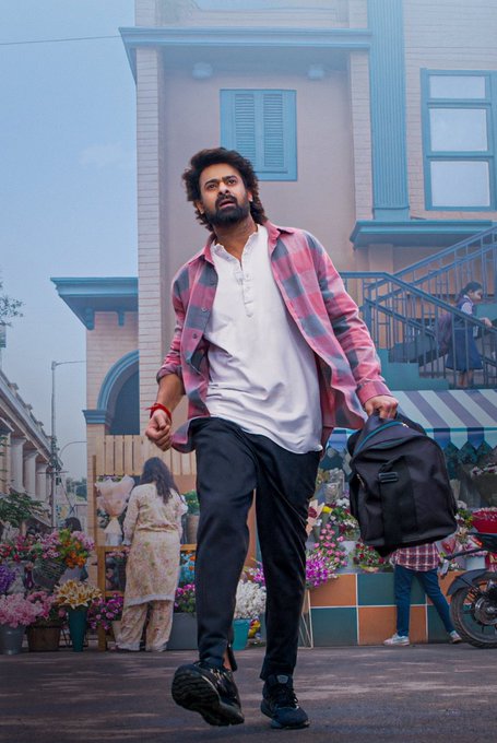 Prabhas wearing a pink and white plaid shirt over a white t-shirt, black pants, and black shoes, carrying a black backpack. He walks on a street with colorful flower stalls, buildings with blue railings, and people in the background.