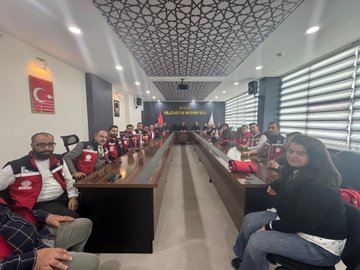 A group of people wearing red vests with "T.C. Milli Eğitim Bakanlığı" text, standing and sitting on steps outside a building with large columns. Another image shows individuals in similar red vests seated around a conference table in a room with Turkish flags, a banner reading "BİTLİS MİLLİ EĞİTİM MÜDÜRLÜĞÜ," and a ceiling with geometric patterns. Metin Tayarer is visible among the group.